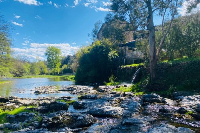 Rivière bordée de rochers et de végétation sous un ciel bleu, avec une maison partiellement visible en arrière-plan.