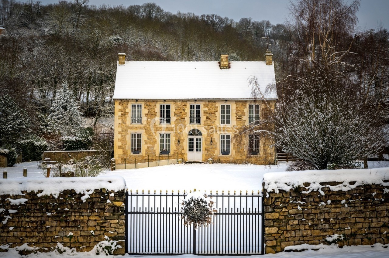 Façade d'une maison en pierre entourée de neige, avec un portail en fer forgé et des arbres en arrière-plan.