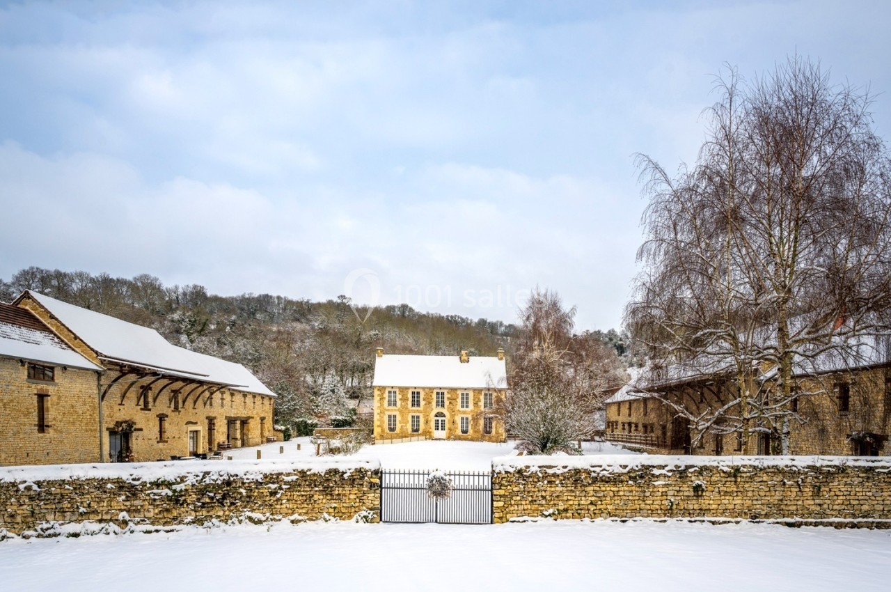 Bâtiments en pierre entourés de neige, avec un portail en fer forgé au premier plan et des arbres dénudés en arrière-plan.