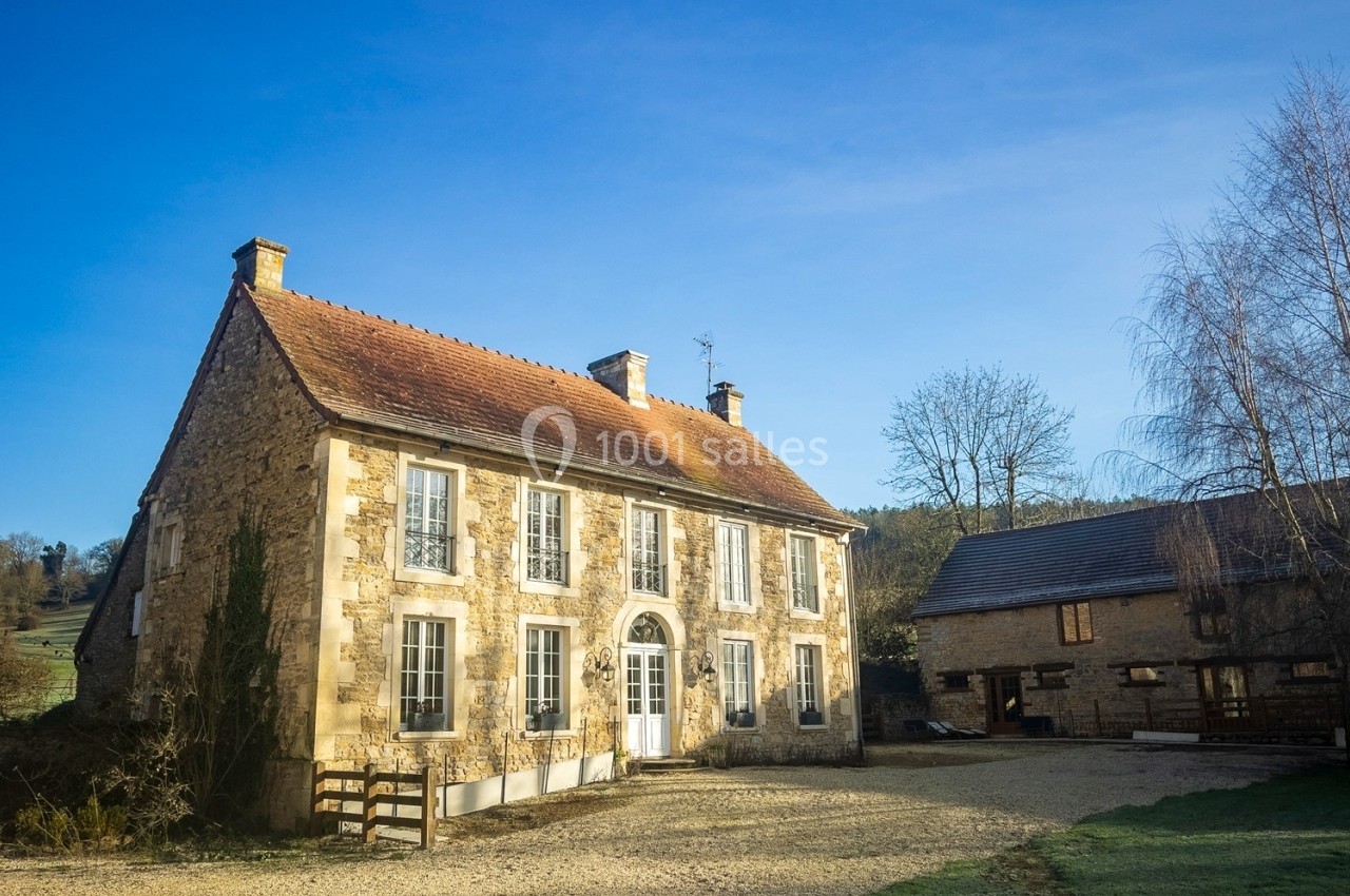 Grande maison en pierre avec volets blancs, entourée d'une cour gravillonnée et de bâtiments annexes, sous un ciel bleu.