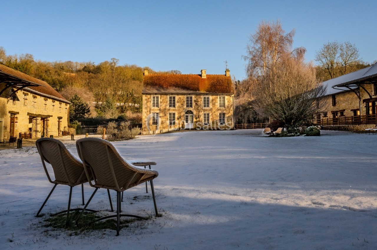 Deux chaises en osier sur une cour enneigée, face à une maison en pierre entourée de bâtiments et d'arbres.
