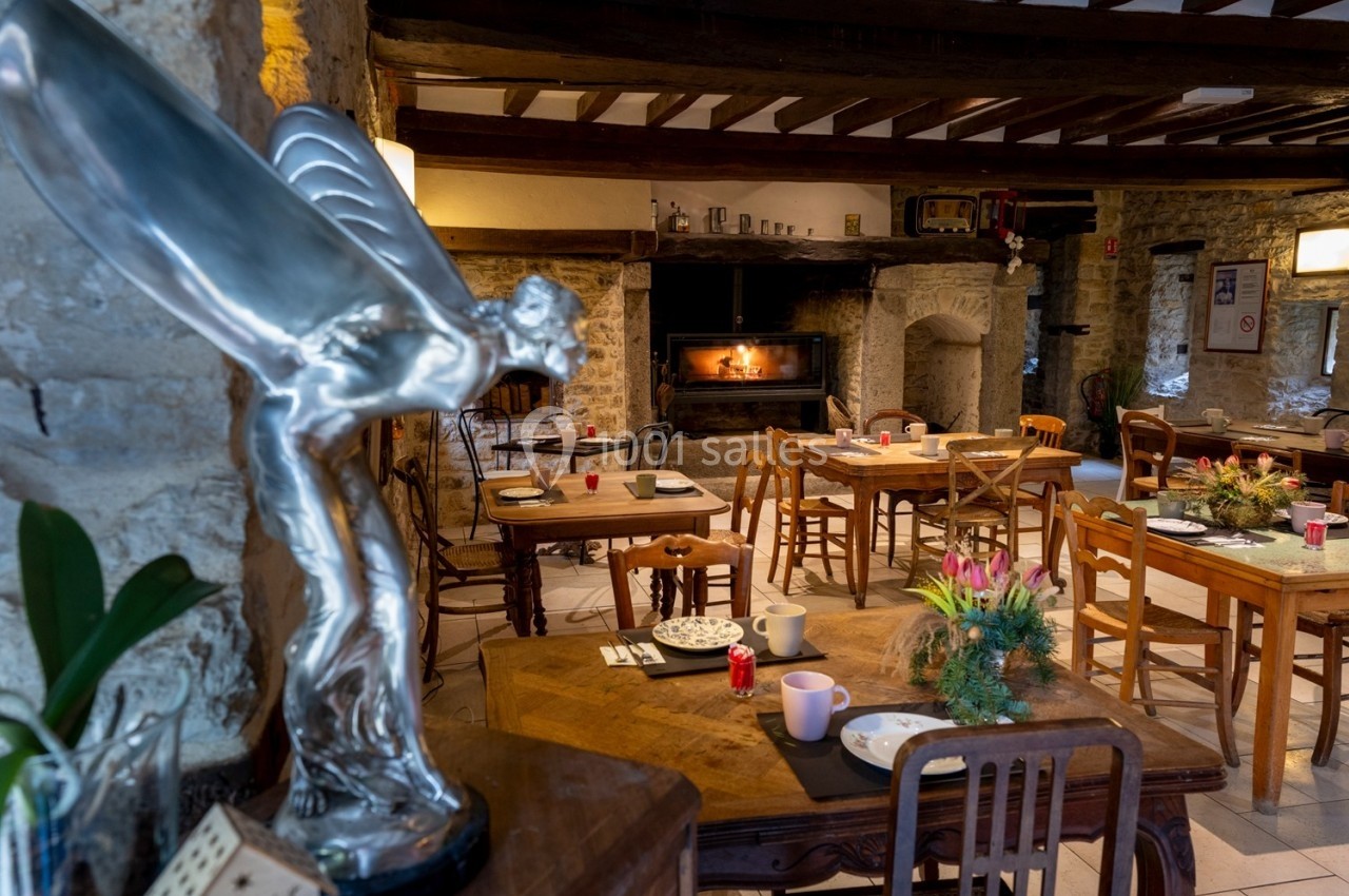 Salle à manger rustique avec cheminée allumée, tables en bois dressées et statue argentée au premier plan.