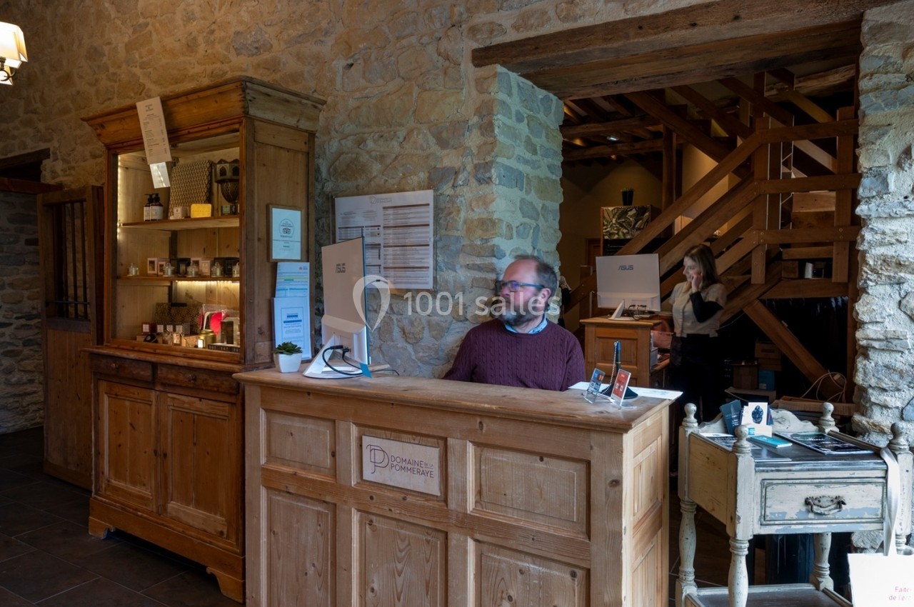 Homme assis à un comptoir en bois dans un espace intérieur en pierre, avec des étagères et des produits exposés.