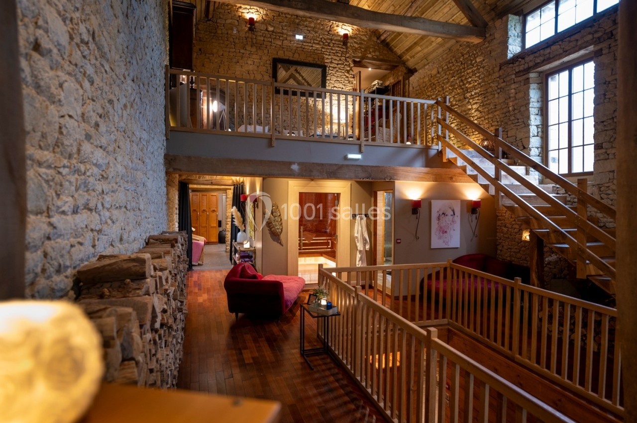 Intérieur d'une maison en pierre avec mezzanine, poutres apparentes, mobilier chaleureux et grande fenêtre lumineuse.
