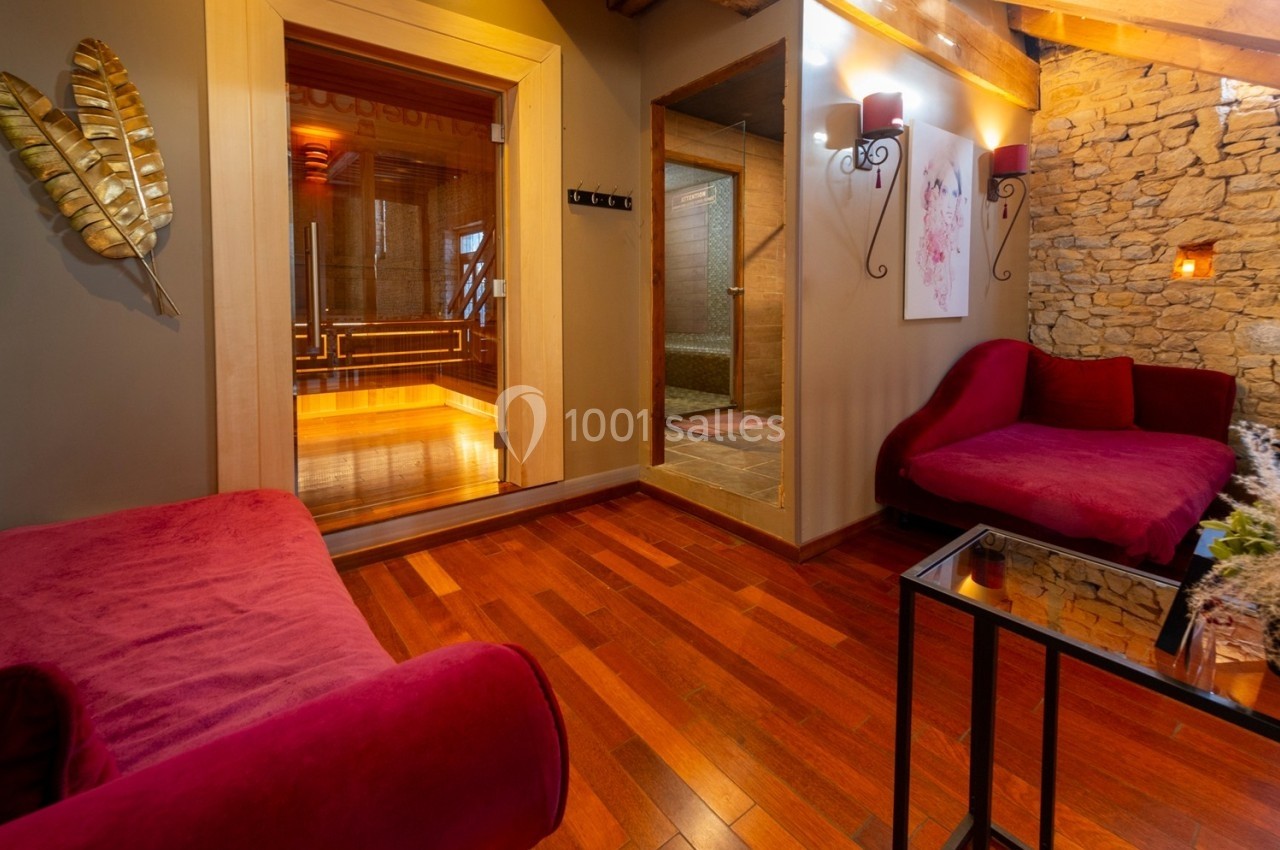 Salon avec fauteuils rouges, parquet en bois, murs en pierre et accès à un sauna vitré éclairé.