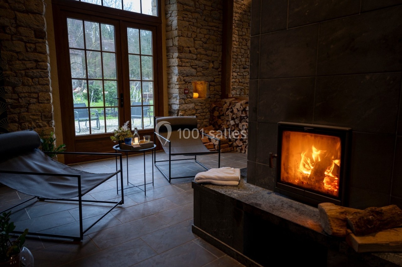 Salon chaleureux avec cheminée allumée, fauteuils modernes, murs en pierre et vue sur un jardin à travers de grandes…