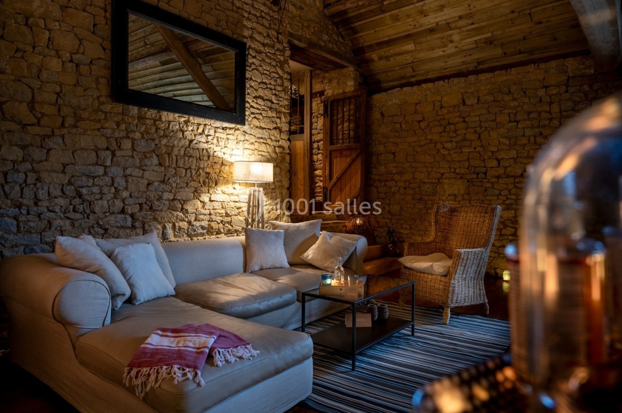 Salon chaleureux avec murs en pierre, canapé beige, fauteuil en osier, éclairage tamisé et décoration cosy.