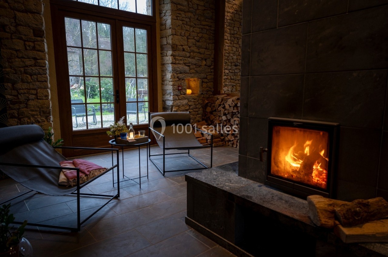 Salon chaleureux avec poêle à bois allumé, fauteuils modernes, murs en pierre et vue sur un jardin à travers de grandes…