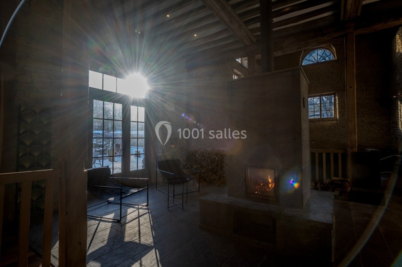 Intérieur d'une pièce avec cheminée allumée, grandes fenêtres laissant entrer la lumière du soleil et murs en briques.
