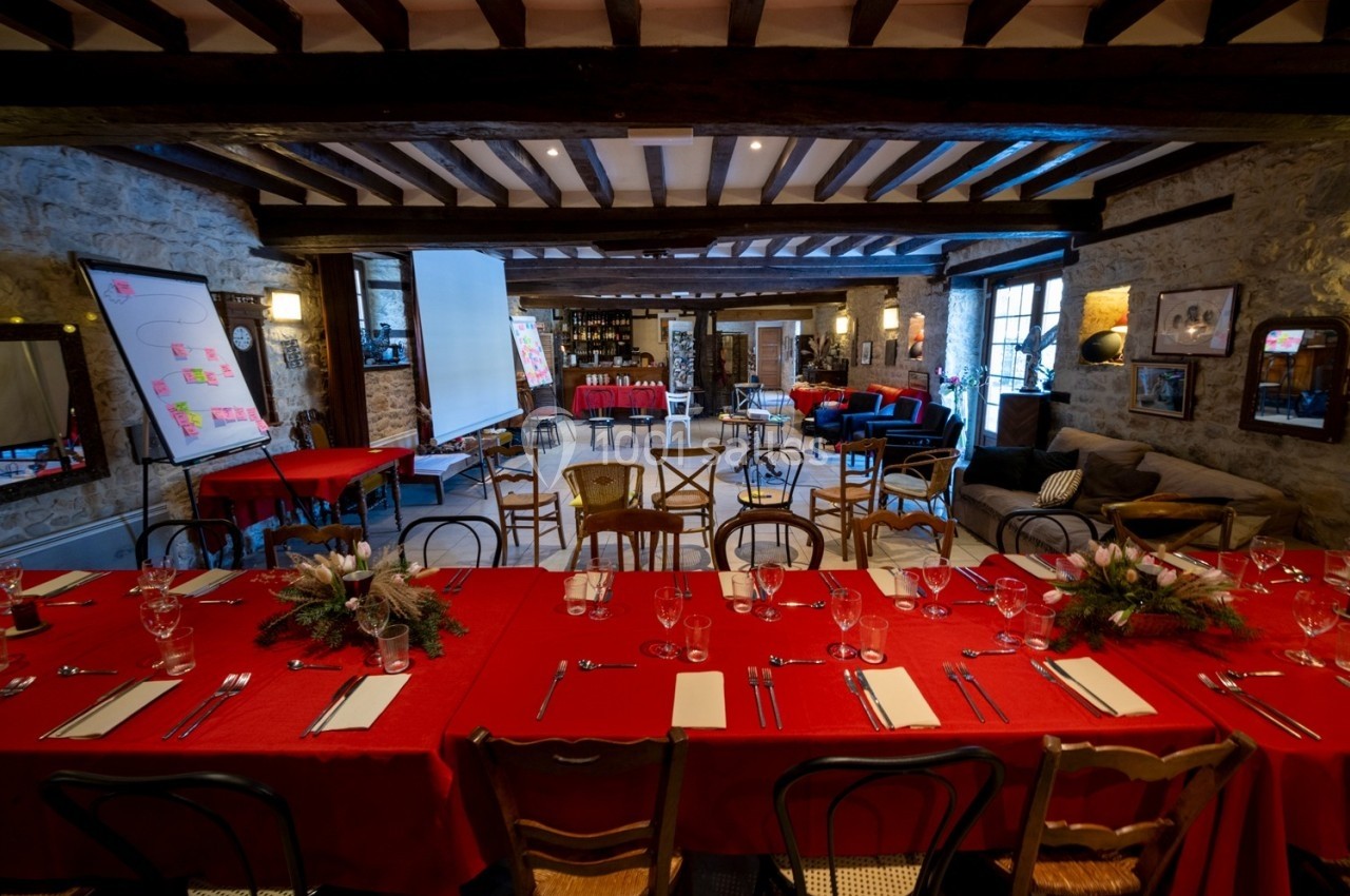 Salle de réception avec tables dressées en rouge, chaises variées, écran de projection et murs en pierre apparente.