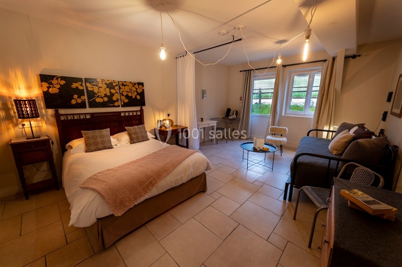 Chambre lumineuse avec lit double, canapé, bureau, table basse et grande fenêtre donnant sur un jardin.