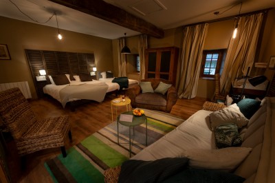 Chambre chaleureuse avec lits, canapé, fauteuils en osier, table basse et éclairage tamisé, décor rustique.