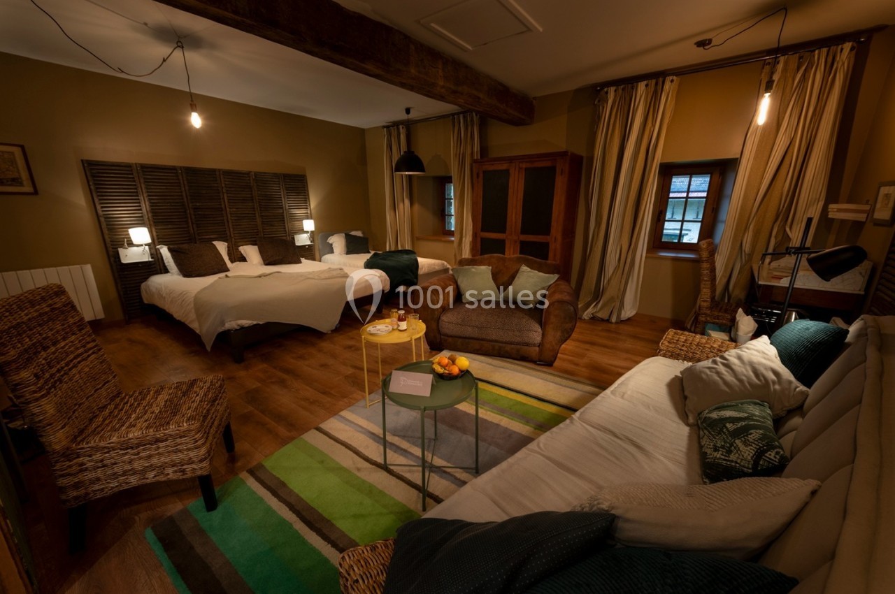 Chambre chaleureuse avec lits, canapé, fauteuils en osier, table basse et éclairage tamisé, décor rustique.