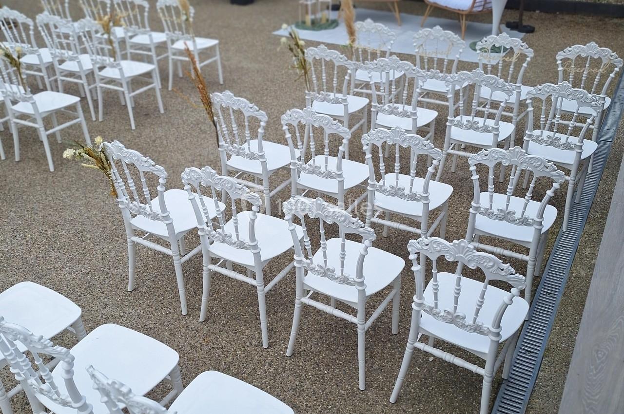 Chaises blanches alignées en extérieur pour une cérémonie, avec deux fauteuils et des décorations florales en arrière-plan.