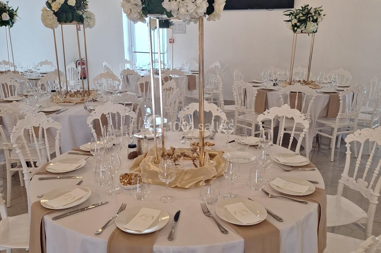 Salle de réception décorée avec des tables rondes, nappes blanches, centres de table fleuris et chaises élégantes.