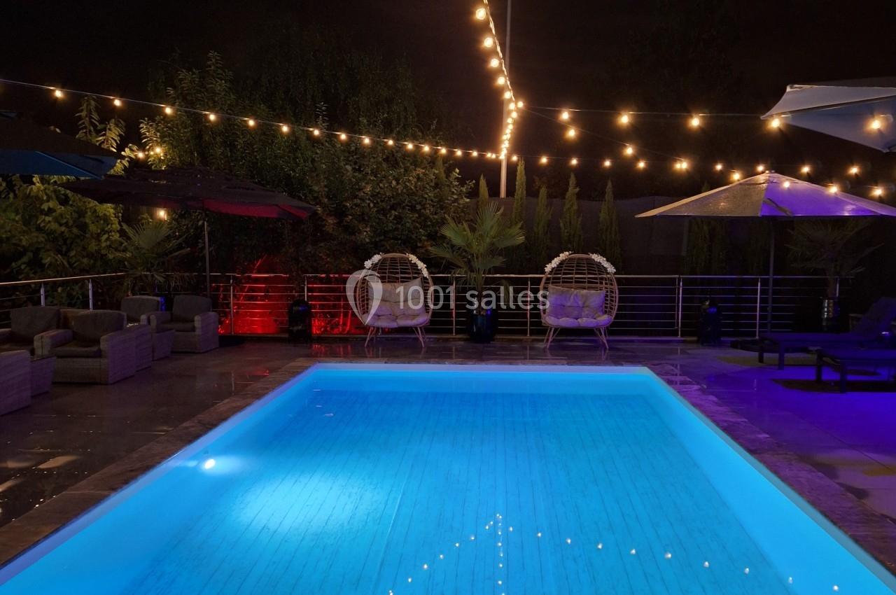 Piscine éclairée de nuit avec guirlandes lumineuses suspendues, entourée de fauteuils et de végétation.