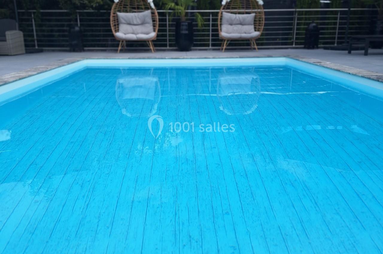Piscine extérieure entourée de fauteuils suspendus, parasols et végétation sous un ciel partiellement nuageux.
