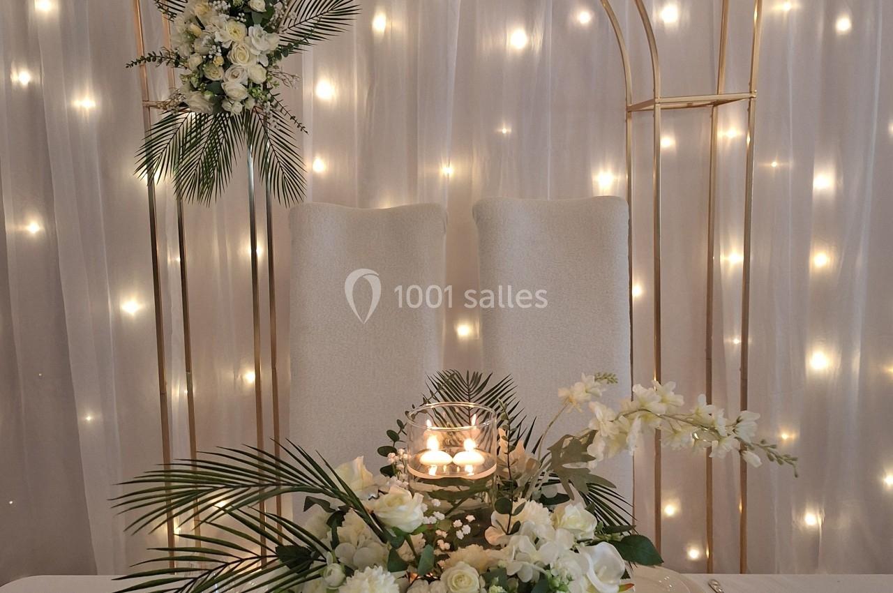 Décoration de table élégante avec fleurs blanches, bougies et arche dorée illuminée par des guirlandes lumineuses.
