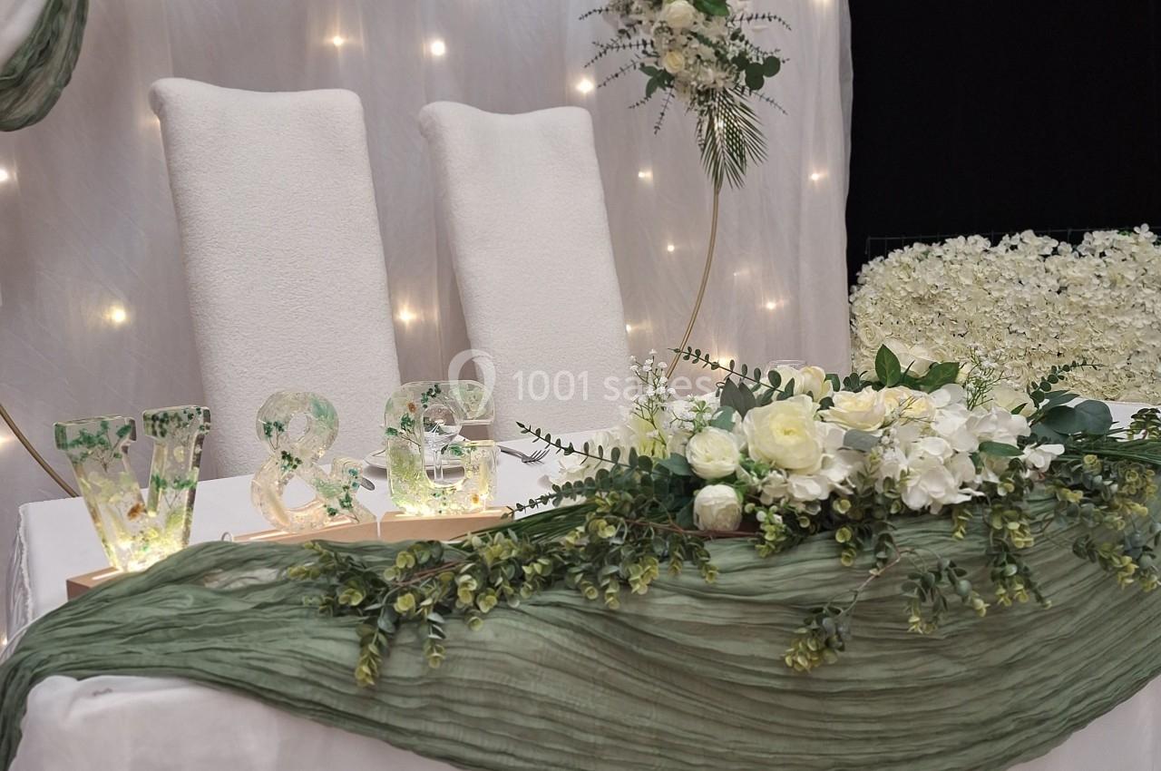 Décoration de table de mariage avec fleurs blanches, feuillage vert, initiales lumineuses et fond éclairé.