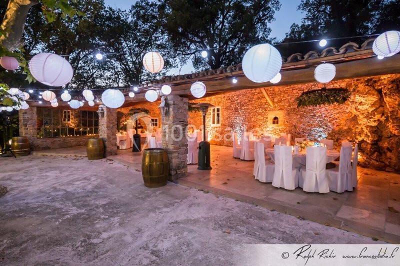 Salle extérieure en pierre éclairée par des guirlandes lumineuses et décorée de lanternes et de tables dressées.