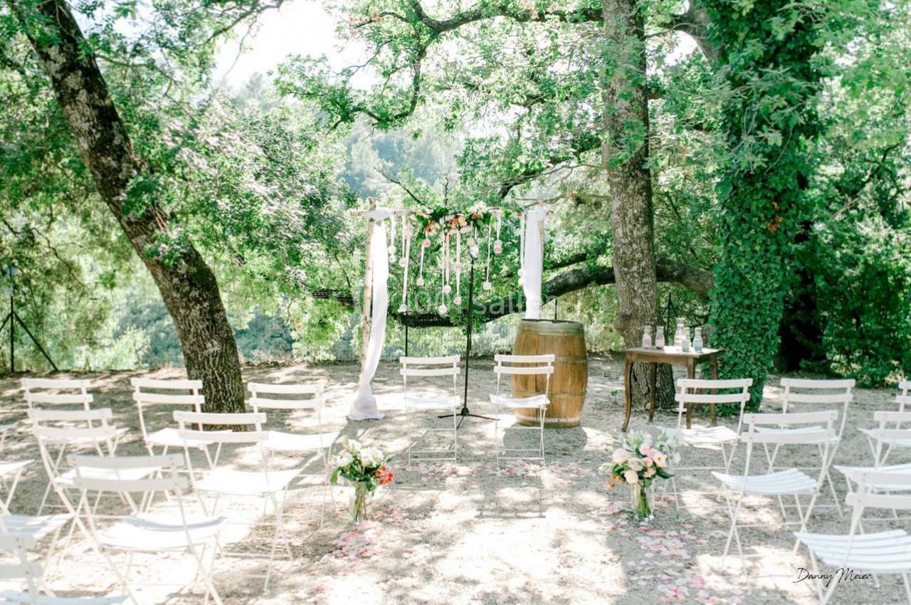 Chaises blanches disposées devant une arche décorée de fleurs et de voilages, dans un cadre boisé en plein air.