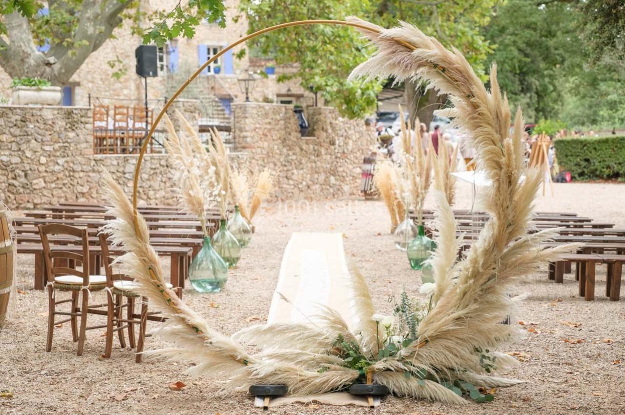Arche circulaire décorée de pampas au centre d'une allée bordée de chaises en bois, dans un cadre extérieur rustique.