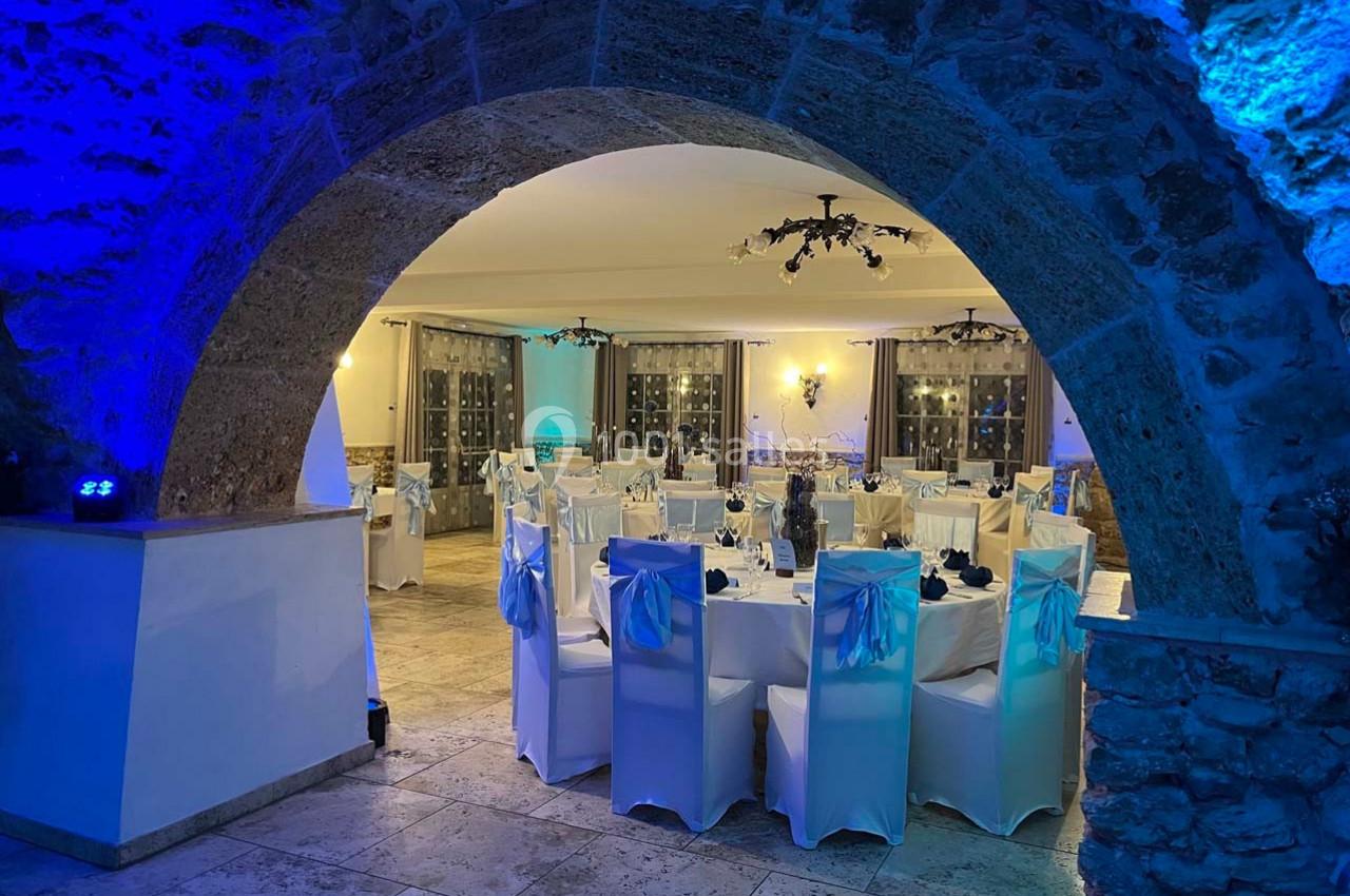 Salle de réception élégamment décorée, avec des tables dressées et des chaises blanches sous un éclairage bleu tamisé.