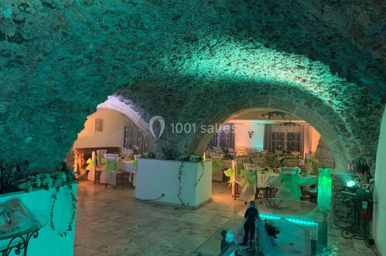 Salle voûtée en pierre avec éclairage vert, tables dressées pour un repas et décoration végétale.