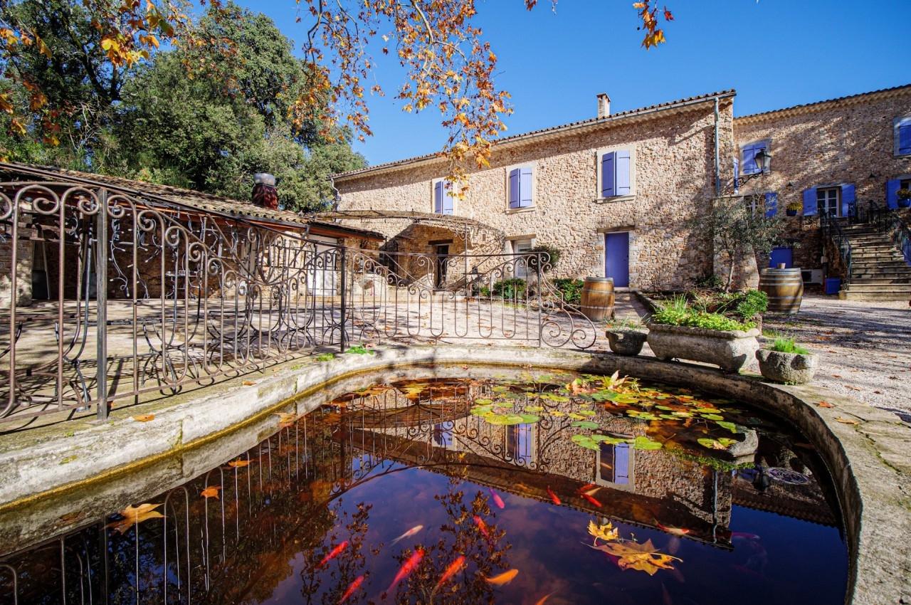 Bassin avec des poissons rouges entouré de feuilles d'automne, devant une bâtisse en pierre sous un ciel dégagé.