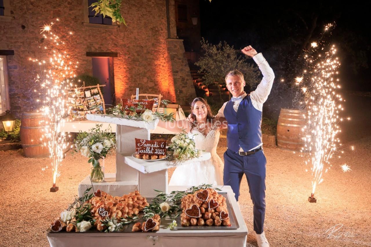 Un couple souriant célèbre leur mariage devant une pièce montée décorée, entourée de feux d'artifice.