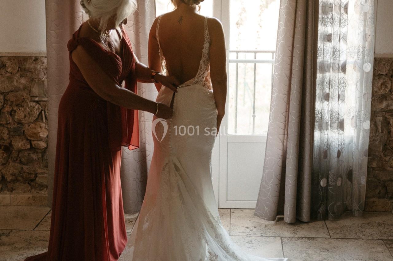 Une femme aide une mariée à ajuster sa robe dans une pièce lumineuse avec des rideaux et une porte-fenêtre en arrière-plan.