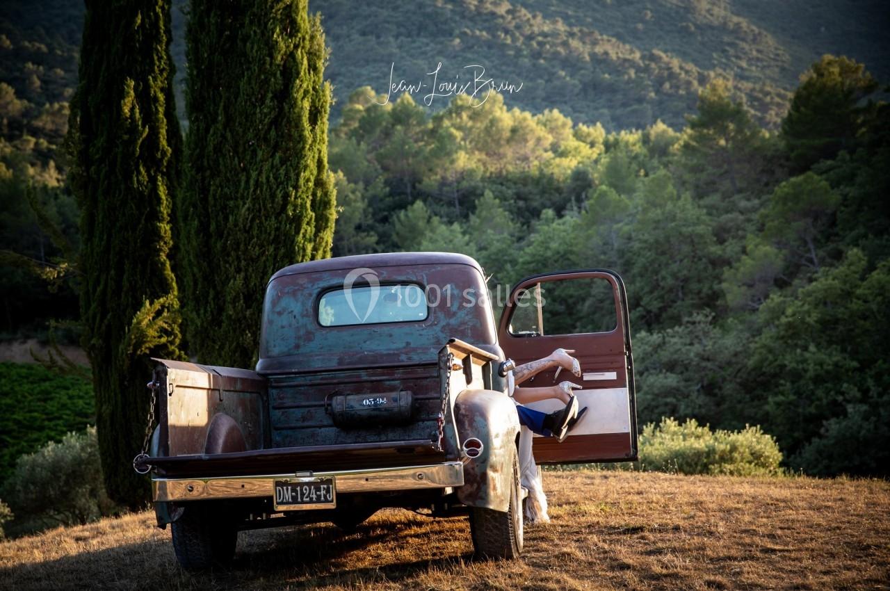 Un vieux pick-up rouillé stationné dans un paysage rural avec des collines, des arbres et une personne assise à l'arrière.