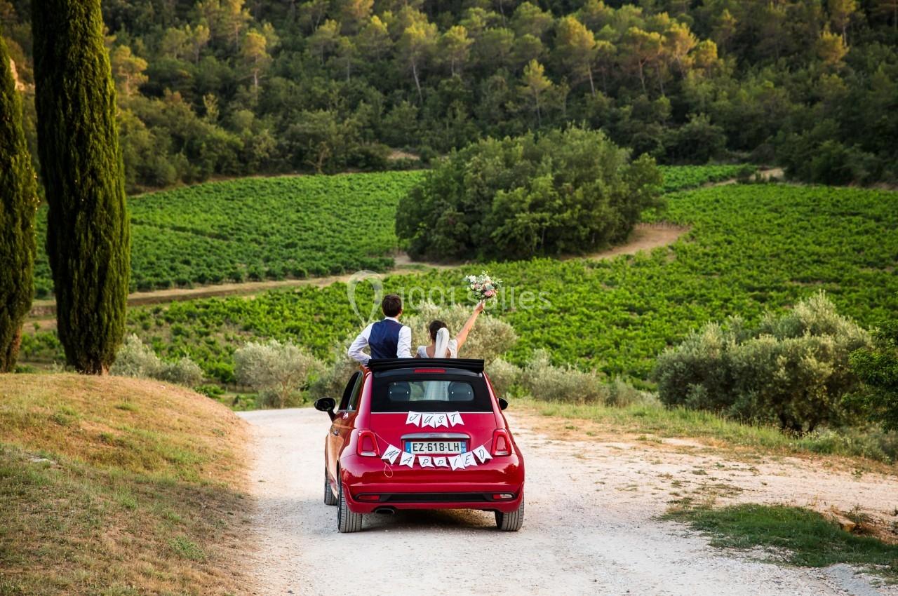 Un couple en tenue de mariage dans une voiture rouge décapotable sur un chemin entouré de vignes et de verdure.