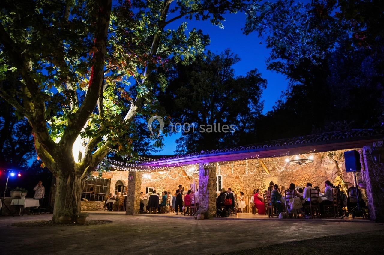 Terrasse éclairée en soirée avec des convives assis sous des guirlandes lumineuses, entourée d'arbres et de nature.