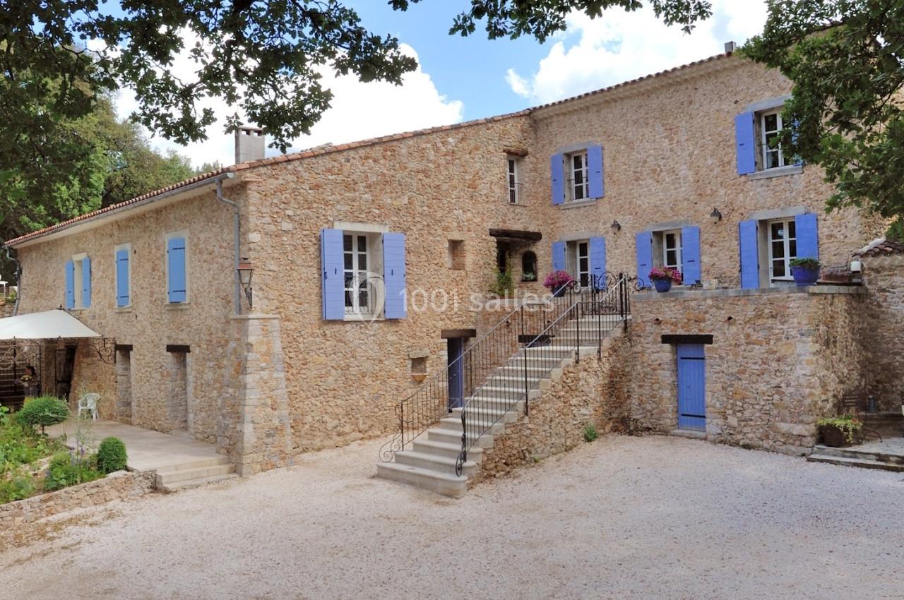 Bâtisse en pierre avec volets bleus, escalier central, cour gravillonnée et arbres en arrière-plan.