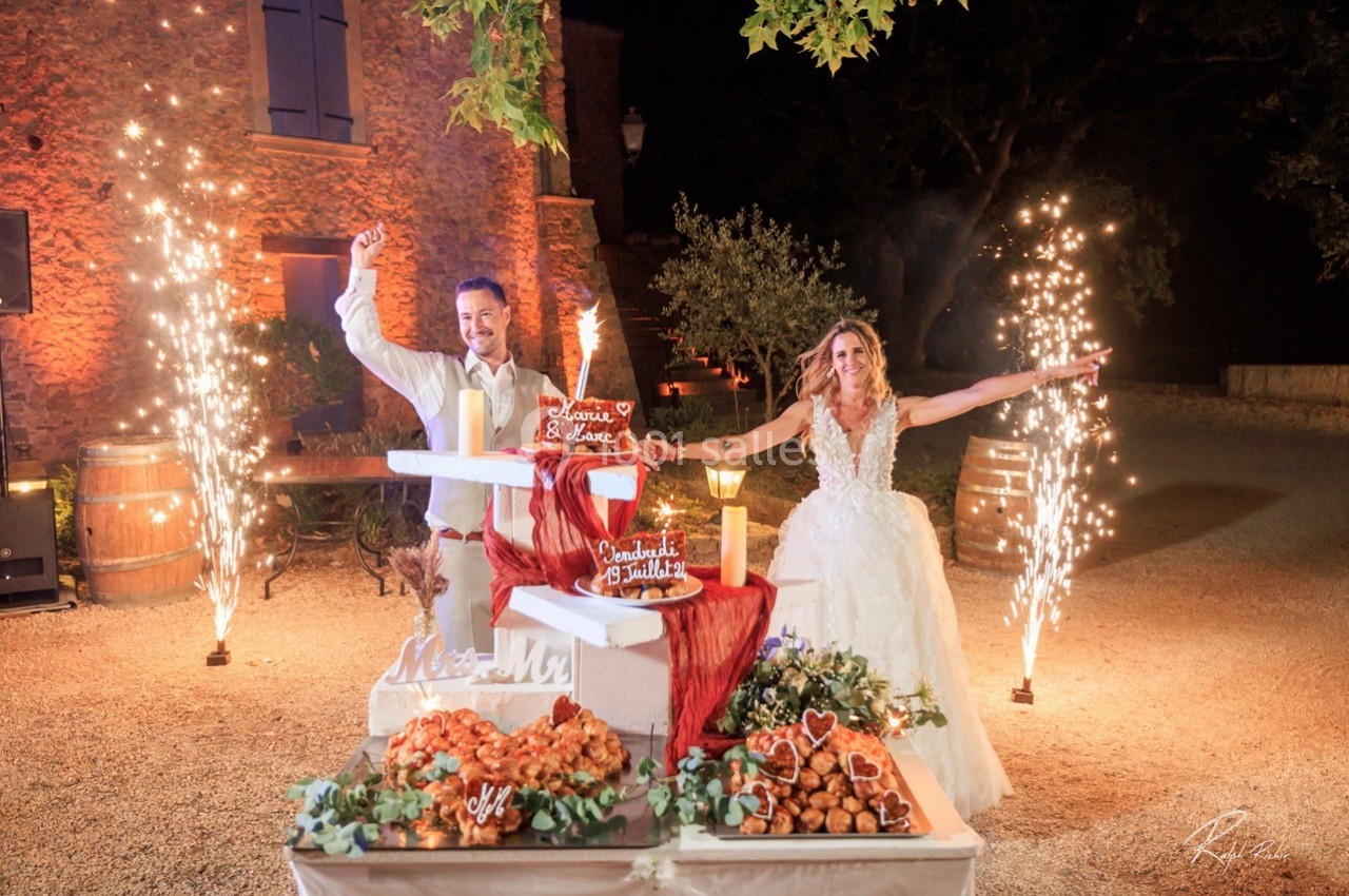 Un couple en tenue de mariage célèbre devant une pièce montée illuminée par des feux d'artifice.