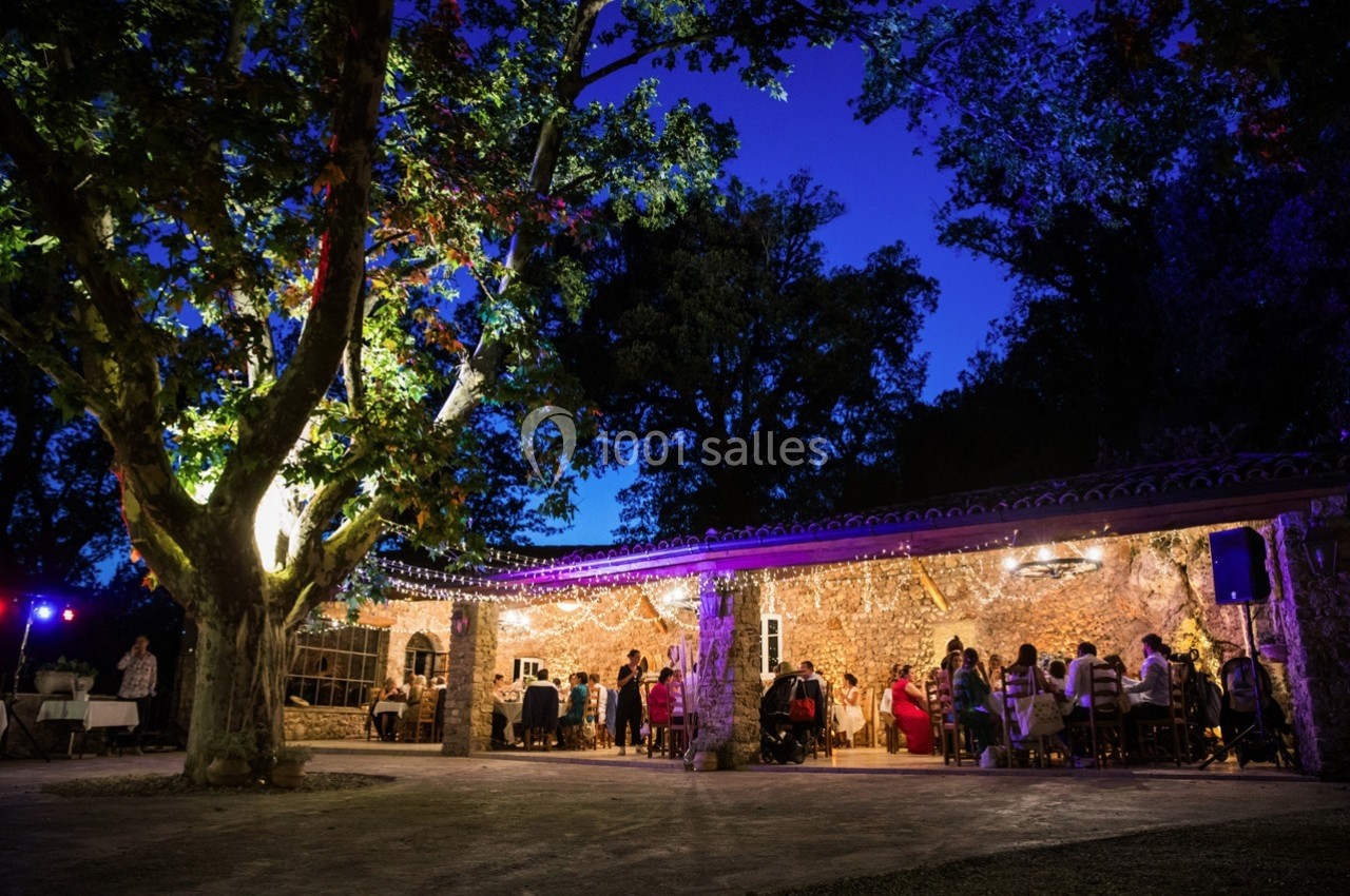 Terrasse éclairée sous un grand arbre, avec des invités assis à des tables lors d'un événement en soirée.