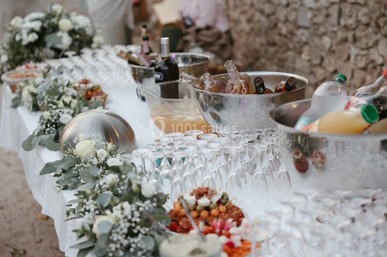 Table de buffet en extérieur avec boissons, verres, fleurs blanches et plats variés disposés sur une nappe blanche.