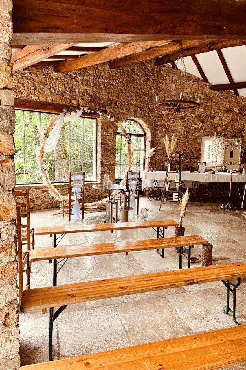 Salle en pierre avec bancs en bois, décor champêtre, grandes fenêtres et table ornée pour un événement.