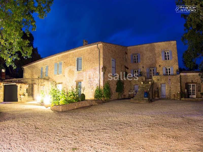 Grande bâtisse en pierre éclairée la nuit, entourée d'une cour en gravier et de végétation.