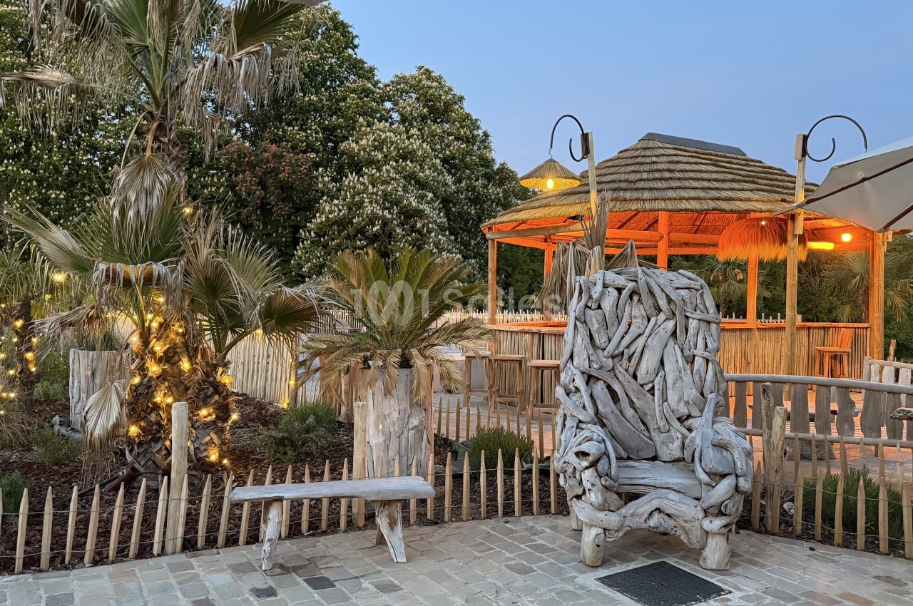 Terrasse extérieure au crépuscule avec un bar en paillote, palmiers illuminés et mobilier en bois flotté.