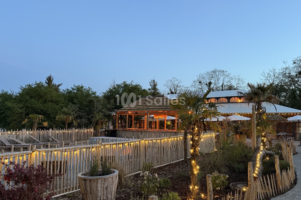 Vue en soirée d'un jardin aménagé de palissades en bois éclairées de guirlandes, avec un kiosque au toit de chaume et une grande tente blanche en arrière-plan. Wait, I must not start with "Vue de". Let me correct: the sentence starts with "Vue en soirée"