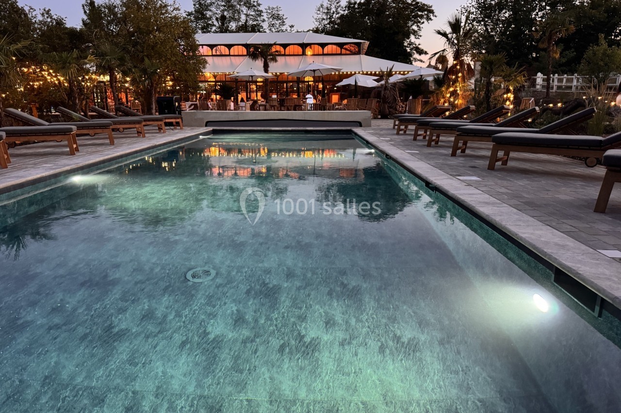 Piscine éclairée au crépuscule avec des transats en bois autour, devant un bâtiment illuminé entouré d'arbres.
