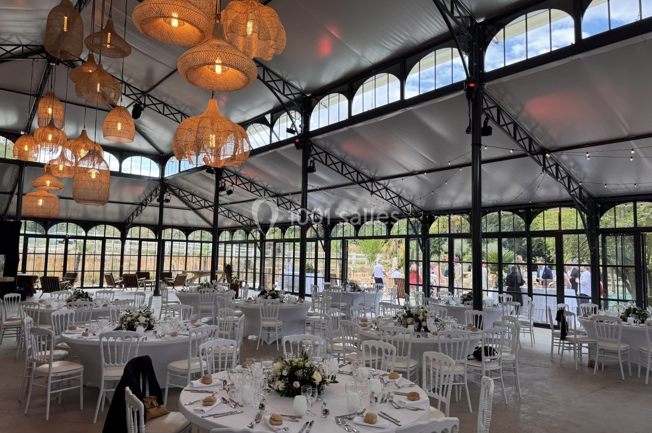 Salle de réception lumineuse avec tables rondes décorées, chaises blanches et suspensions en osier sous une verrière.