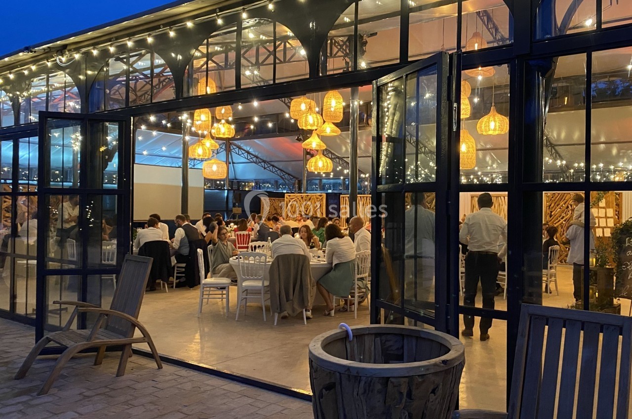 Salle de réception vitrée éclairée par des guirlandes et des suspensions, avec des invités assis à table le soir.