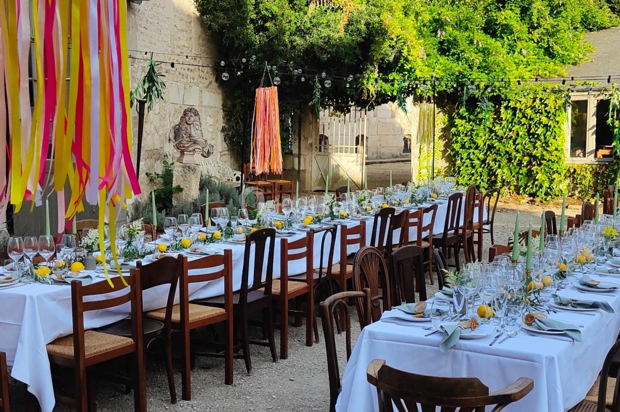 Tables dressées avec nappes blanches, chaises en bois et décorations colorées dans une cour extérieure ensoleillée.