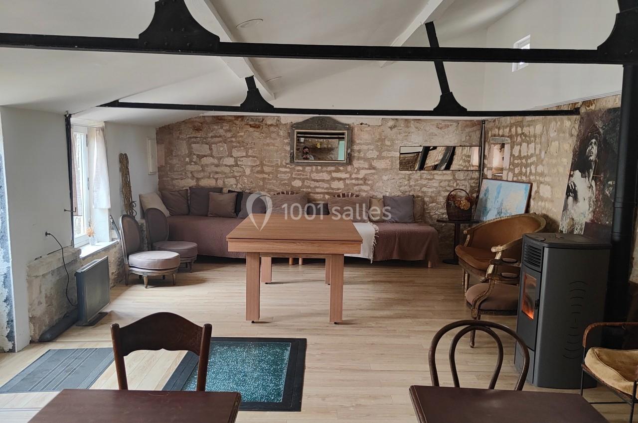 Salle lumineuse avec murs en pierre, tables en bois, poêle à bois et canapé dans un espace chaleureux.