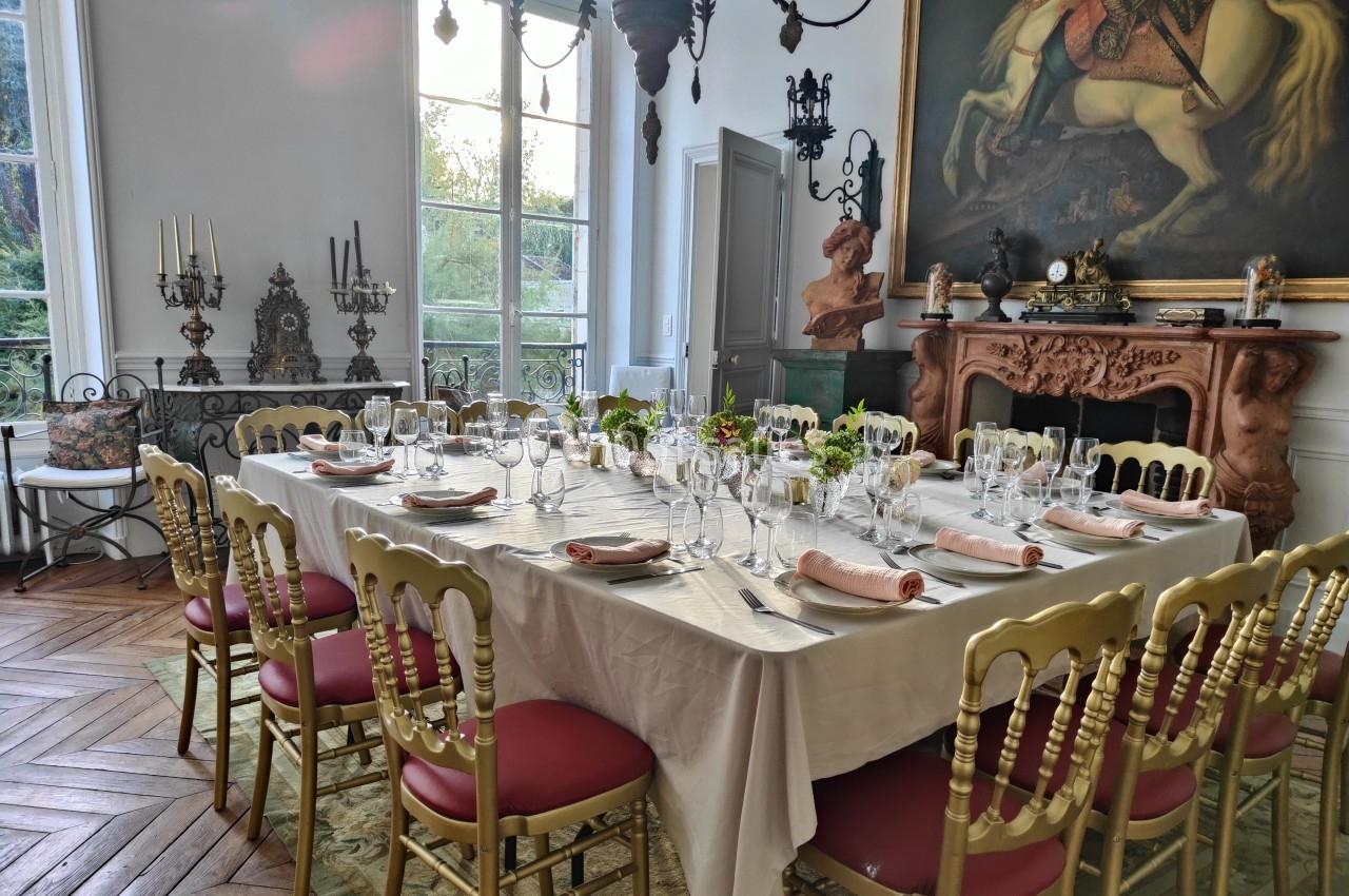 Table dressée pour un repas dans une salle élégante avec cheminée, grandes fenêtres et décorations classiques.