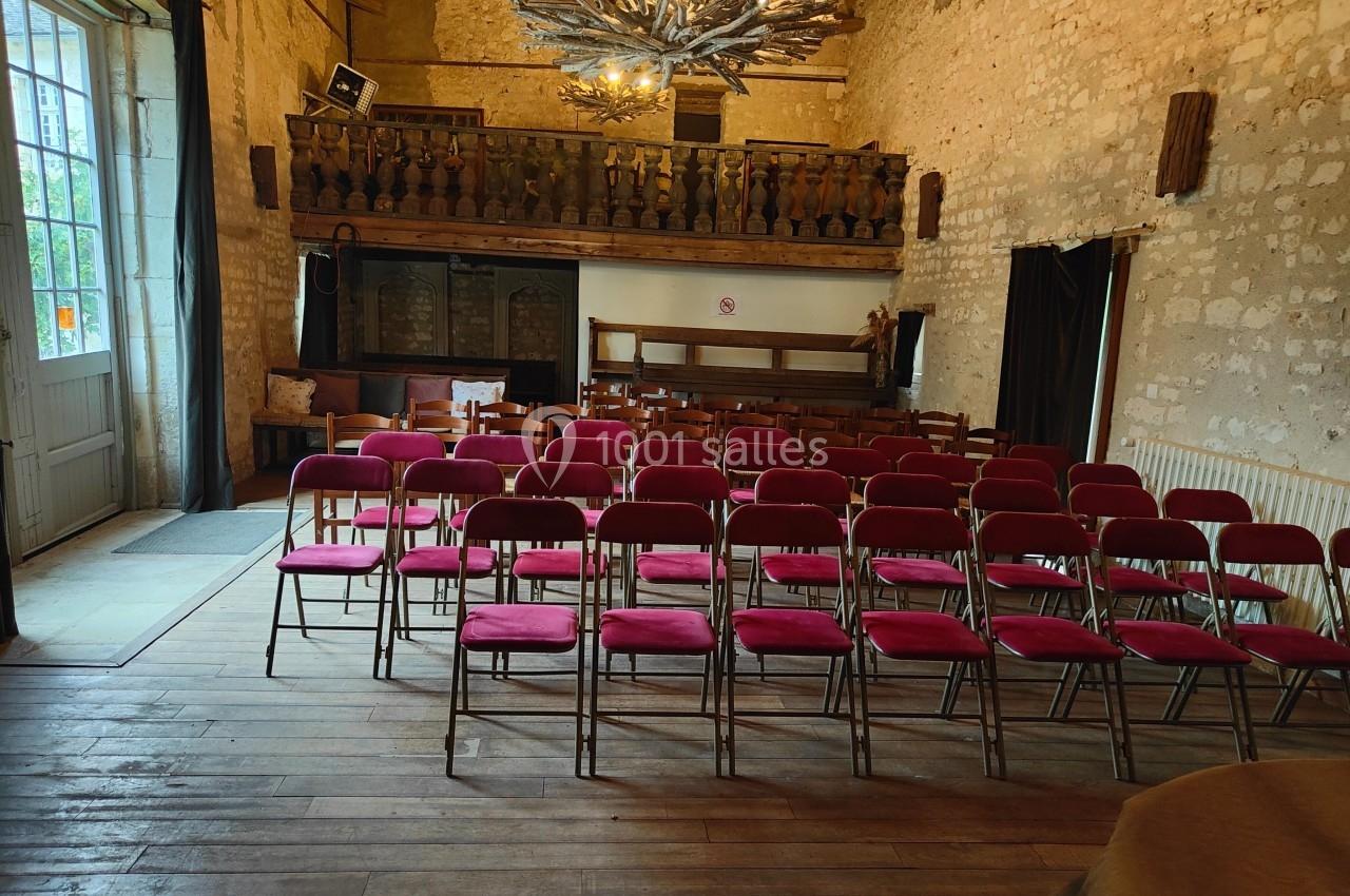 Salle en pierre avec des rangées de chaises rouges alignées face à une petite scène en bois, éclairée par une suspension.