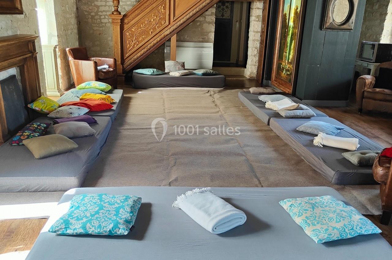 Matelas et coussins disposés au sol dans une pièce rustique avec poutres apparentes et escalier en bois sculpté.